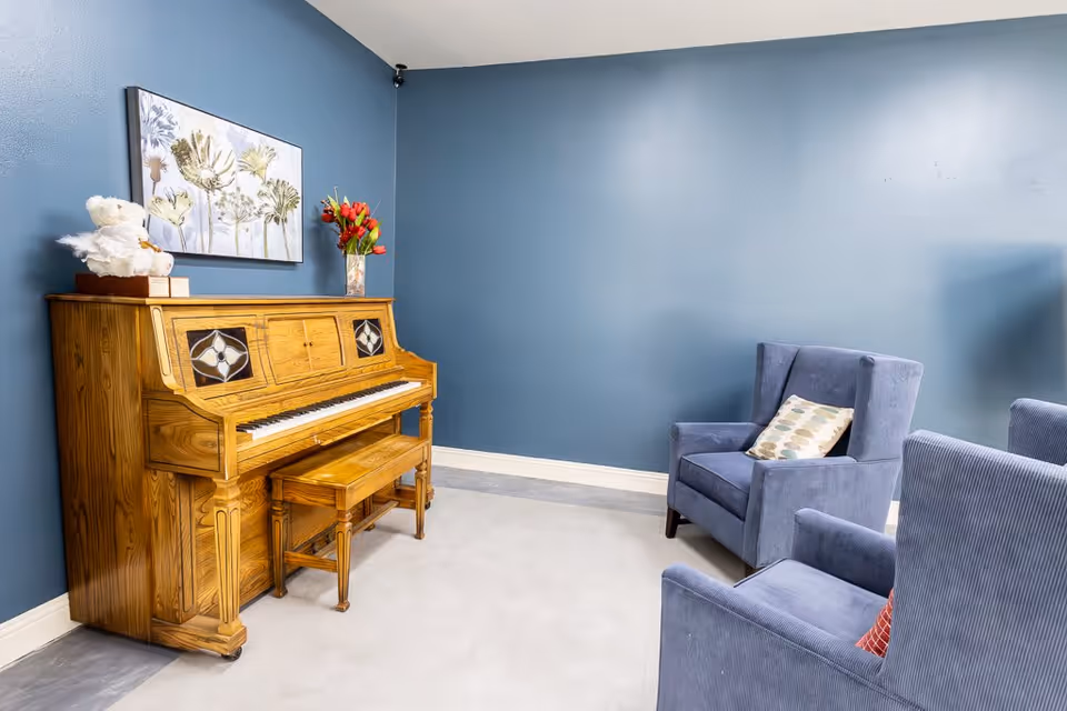 A cozy room with blue walls featuring a wooden upright piano with a matching bench. On top of the piano, there is a white teddy bear and a vase with red flowers. Two blue upholstered armchairs with patterned cushions are positioned facing the piano. A floral painting hangs above the piano.