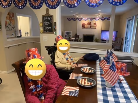 A festive dining room decorated with patriotic American flags and red, white, and blue bunting. Two people with smiley face emojis covering their faces sit at a table set with plates of food arranged in the American flag pattern. The room has a cozy atmosphere with a fireplace and a television in the background.