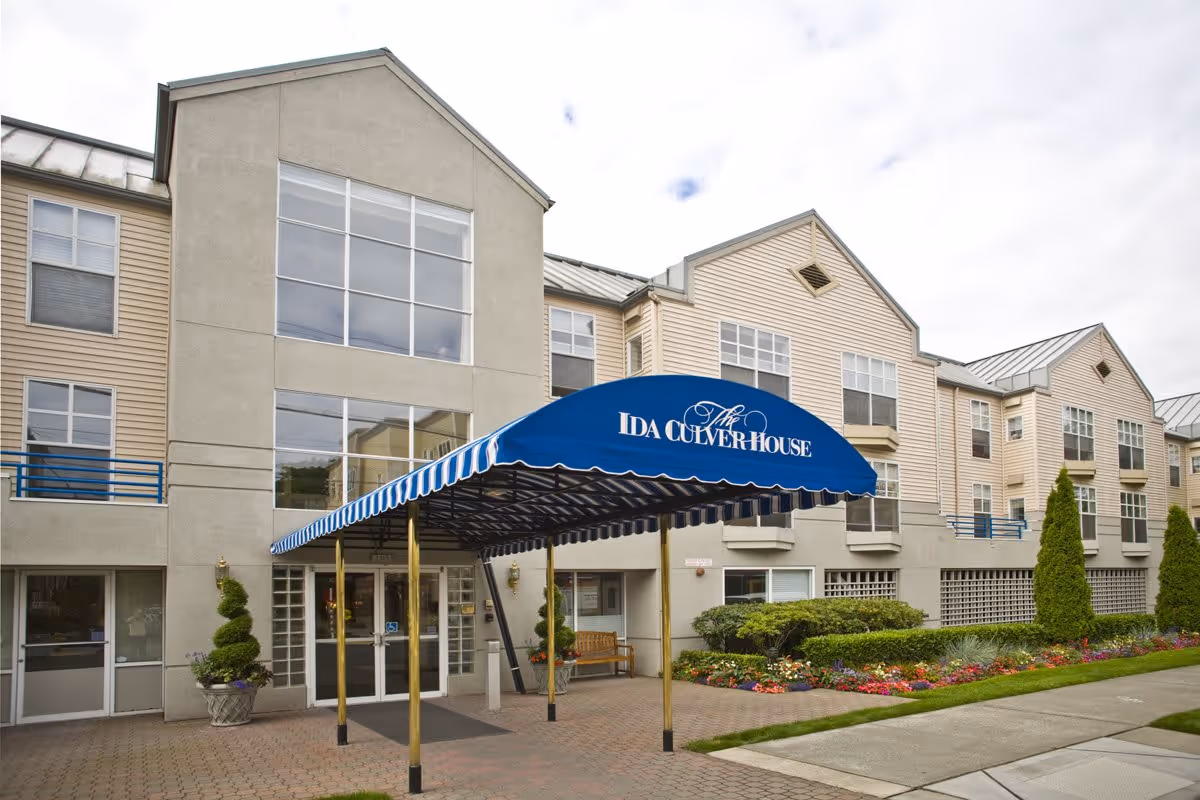 Exterior view of Ida Culver House Ravenna showing the entrance with a blue canopy that has the facility's name on it. The building is beige with large windows and landscaped flower beds along the walkway.