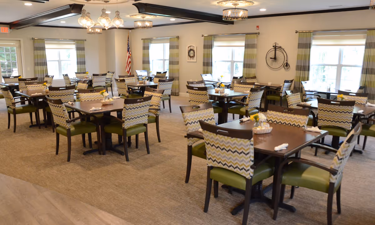 A spacious dining room with multiple square wooden tables and cushioned chairs featuring a zigzag patterned backrest and green seats. The room has large windows with striped curtains, beige carpet flooring, and decorative light fixtures hanging from the ceiling. An American flag is visible in the corner, and the walls are adorned with minimal decor including a vintage bicycle wall clock.