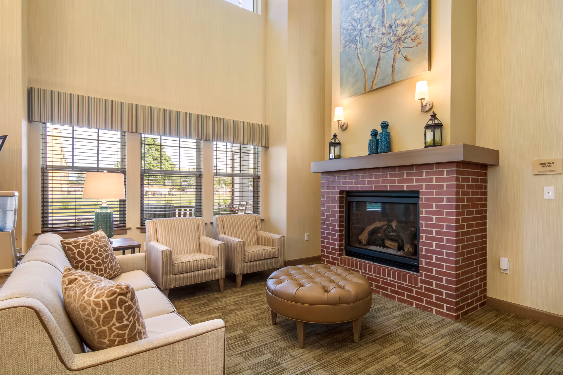 A cozy living room area with a beige sofa adorned with patterned pillows, two striped armchairs, and a round tufted leather ottoman in front of a brick fireplace. The fireplace mantel is decorated with two lanterns and three blue vases. Above the mantel hangs a large floral painting. The room has tall windows with blinds and a striped valance, allowing natural light to brighten the space. A table lamp is placed beside the sofa, and a sign on the wall indicates a meeting room nearby.