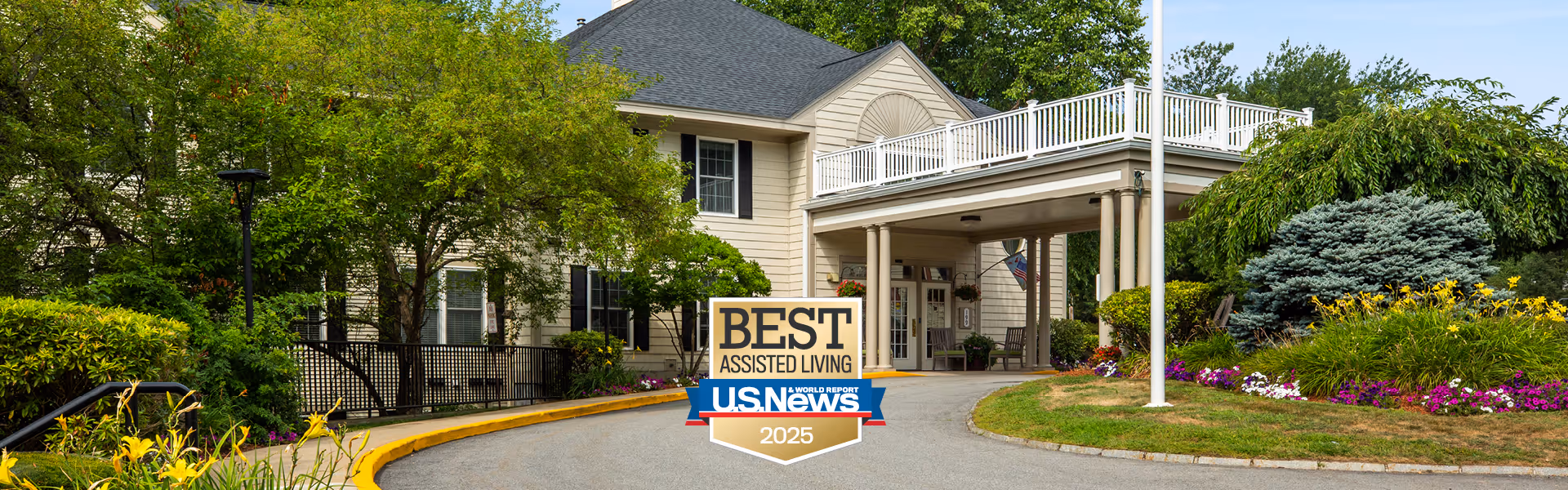 Exterior view of a senior living facility with a driveway leading to a covered entrance. The building is surrounded by well-maintained greenery, including trees, bushes, and colorful flowers. A sign in front of the building reads 'BEST ASSISTED LIVING U.S. News 2025'.