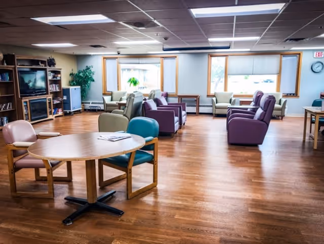 A spacious senior living common area with wooden flooring, multiple armchairs in beige and purple arranged in small groups, a round wooden table with two chairs in the foreground, large windows letting in natural light, a TV on a wooden stand, bookshelves, and a potted plant in the corner.