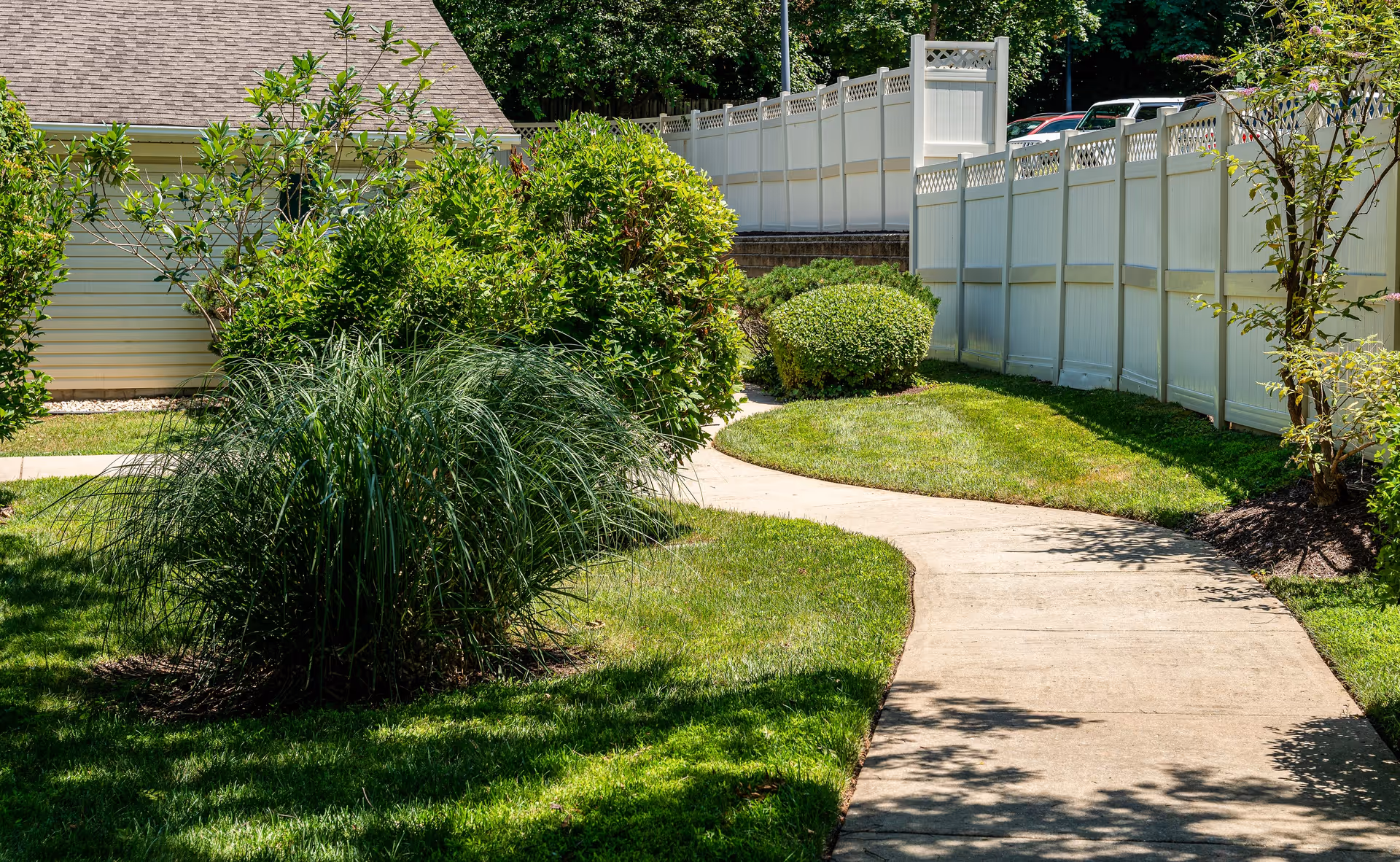 A winding concrete pathway through a well-maintained garden area with green grass, bushes, and small trees. A white fence runs along the right side of the path, and part of a building with beige siding and a shingled roof is visible on the left.