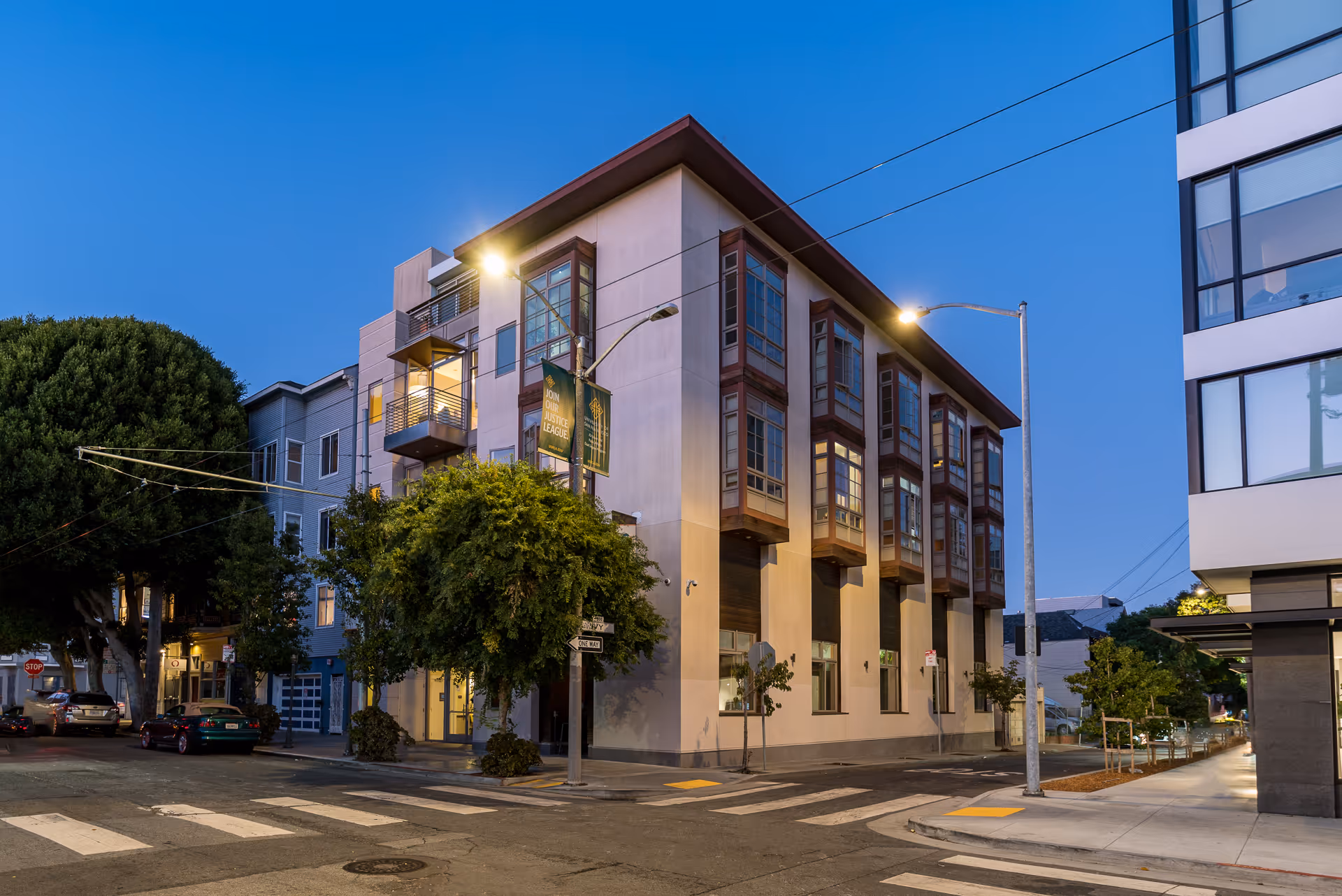 Street view of a modern multi-story building at dusk with street lamps on, trees, parked cars, and a clear sky.