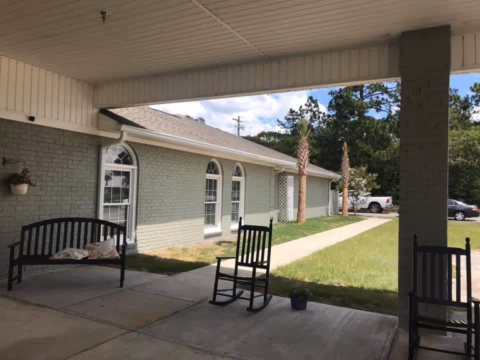 Covered entrance porch with rocking chairs and a bench facing a walkway beside a light-green brick building with arched windows and palm trees.