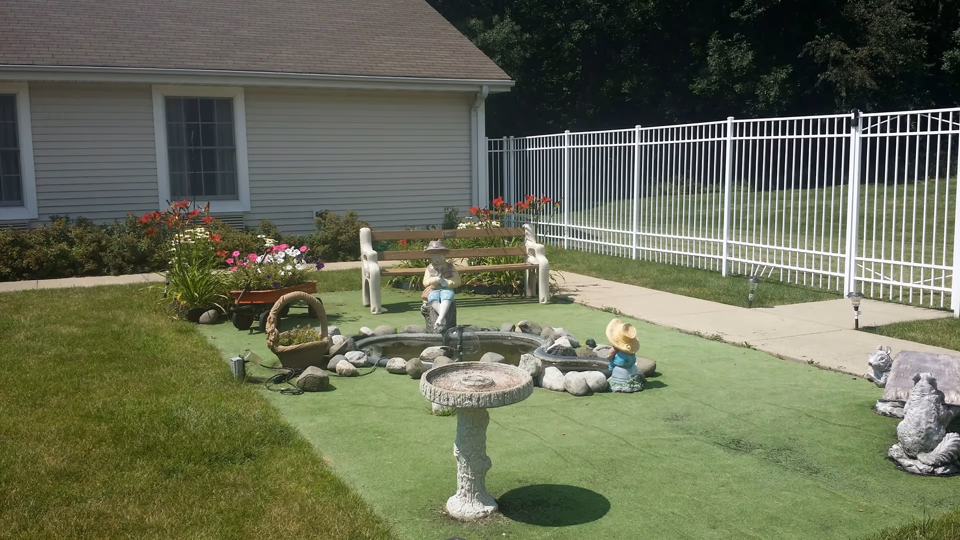 Outdoor garden area with a small circular pond surrounded by rocks and garden statues, including a figure wearing a hat. There is a wooden bench behind the pond, flower beds with colorful flowers, a white metal fence, and a building with beige siding and windows in the background.