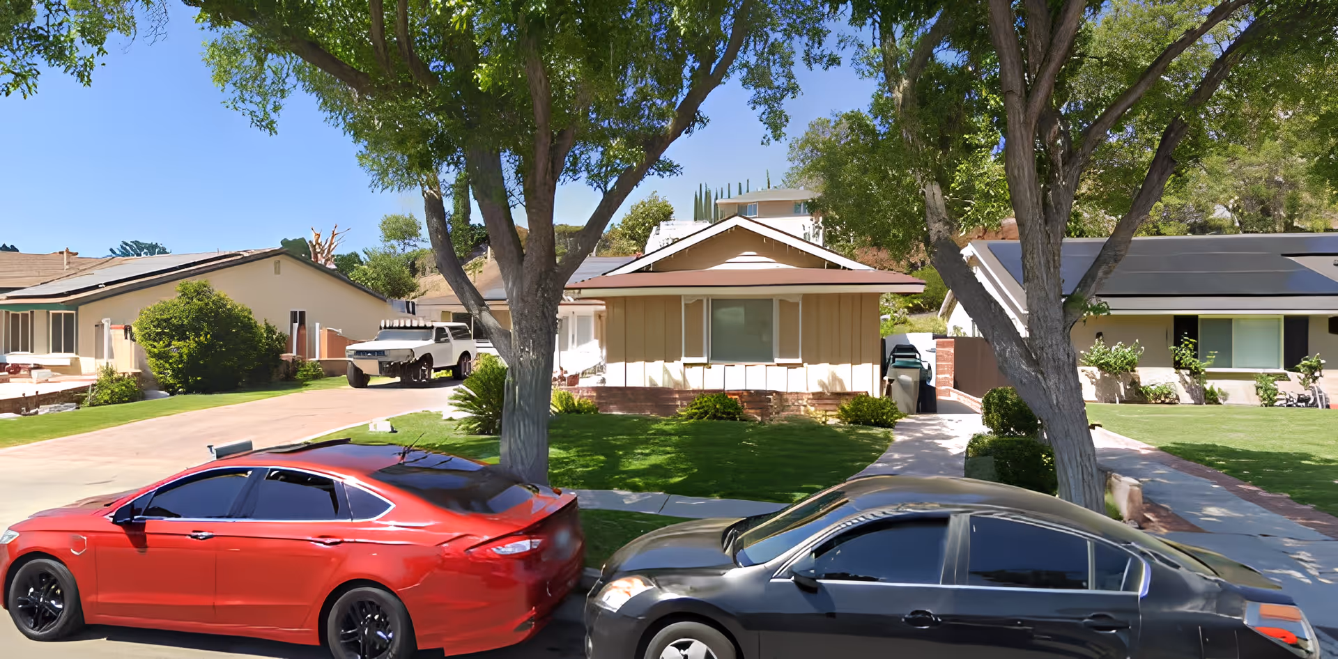 View of a residential street with single-story houses, green lawns, and large trees. Two cars, one red and one black, are parked on the street in front of the houses. The sky is clear and blue.