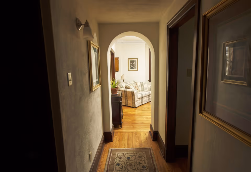 View down a narrow hallway with wooden floors and light-colored walls, leading to a living room area with a patterned sofa, a framed picture on the wall, and a small table with a plant and lamp.