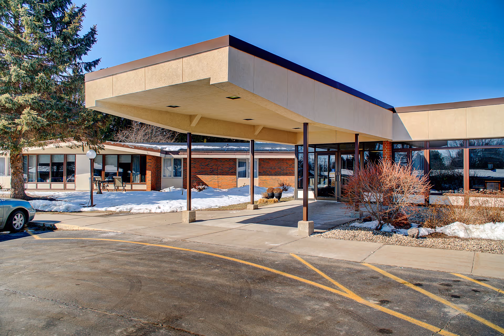 Exterior view of Evansville Manor Skilled Nursing facility entrance with a covered drop-off area, brick and beige walls, large windows, some snow on the ground, and a tree on the left side.