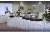 A buffet setup in a dining area with tables covered in white tablecloths, chafing dishes, floral centerpieces, and glassware arranged neatly. The room has framed artwork on the walls and large windows with blinds.