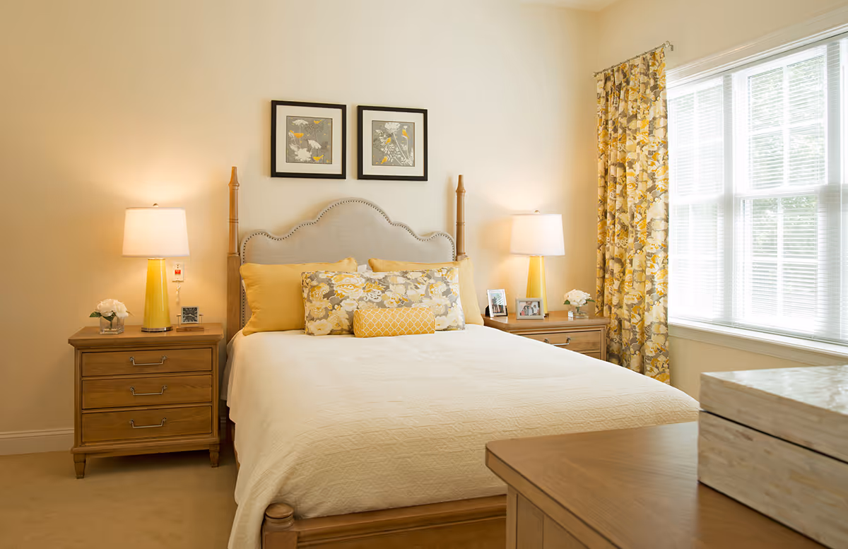 A cozy bedroom with a wooden bed frame and a beige upholstered headboard. The bed is made with a cream-colored quilt and decorated with yellow and floral pillows. On each side of the bed are wooden nightstands with yellow lamps, small flower arrangements, and framed photos. A large window with floral yellow curtains allows natural light to fill the room.