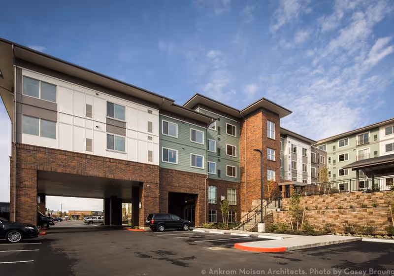 Exterior view of Merrill Gardens at Cedar Mill, a multi-story senior living facility with a combination of brick and light-colored siding. The building features multiple windows, a covered parking area, and a landscaped area with stairs leading up to an entrance. The sky is partly cloudy.
