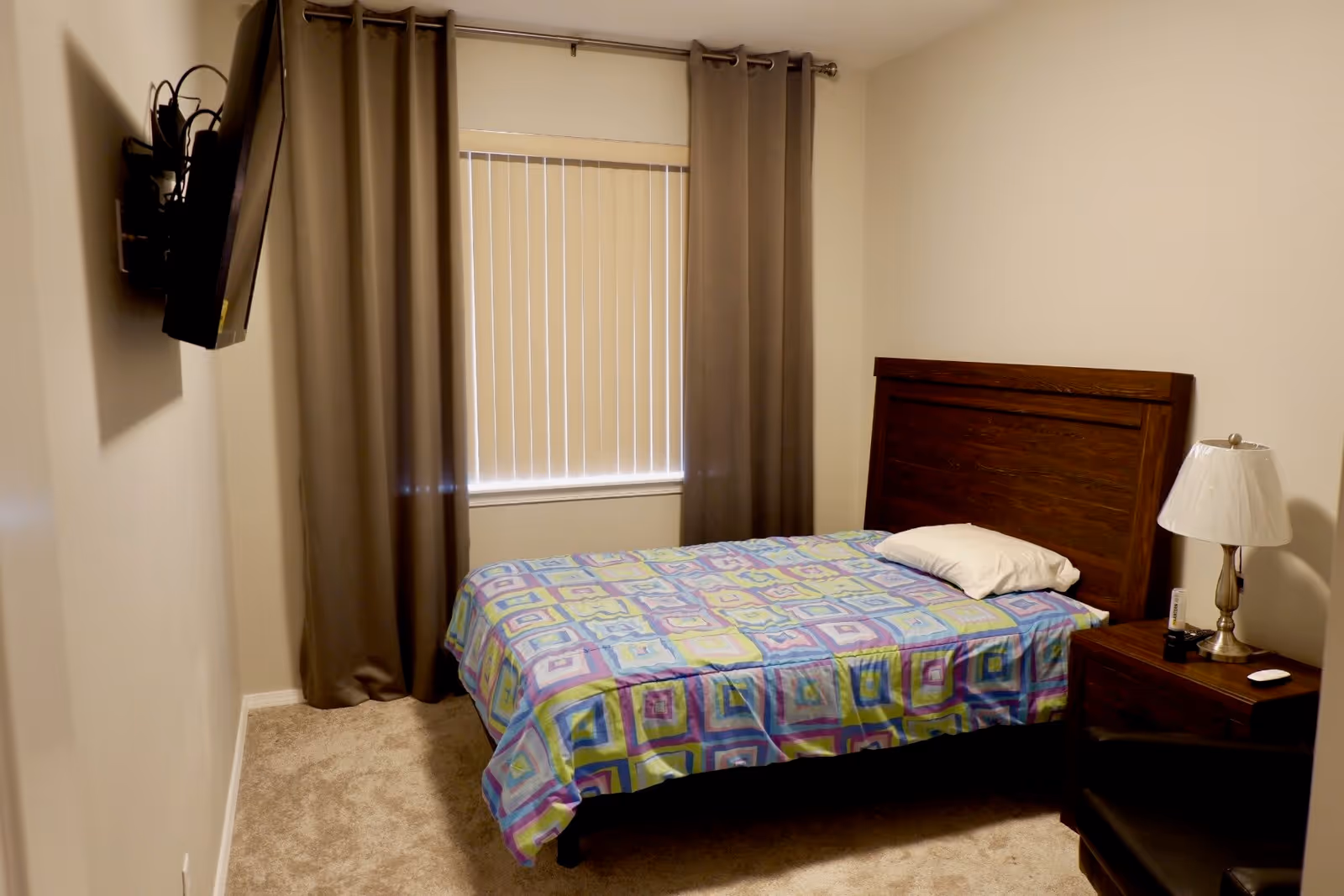 Small bedroom with a single bed and wooden headboard, colorful quilt, nightstand with a lamp, wall-mounted TV, and a window with blinds and curtains.