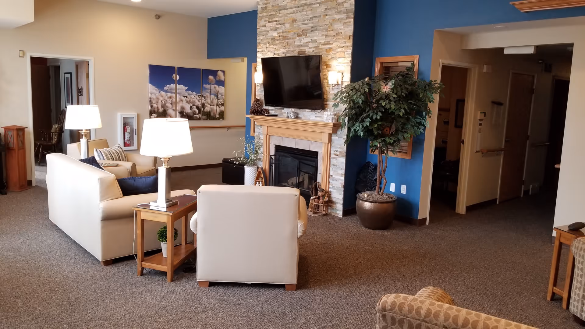 A cozy living room area in Timberwood Lodge Assisted Living featuring beige sofas and armchairs arranged around a stone fireplace with a mounted flat-screen TV. The room has a blue accent wall, a large potted plant, side tables with lamps, and a triptych wall art depicting cotton plants. The carpeted floor and warm lighting create a welcoming atmosphere.