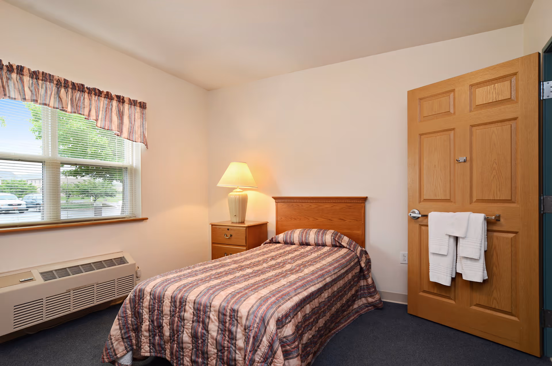 A small bedroom with a single bed covered in a striped quilt. Next to the bed is a wooden nightstand with a lamp. A window with blinds and a valance is on the left wall, and a wooden door with two white towels hanging on a towel rack is on the right.
