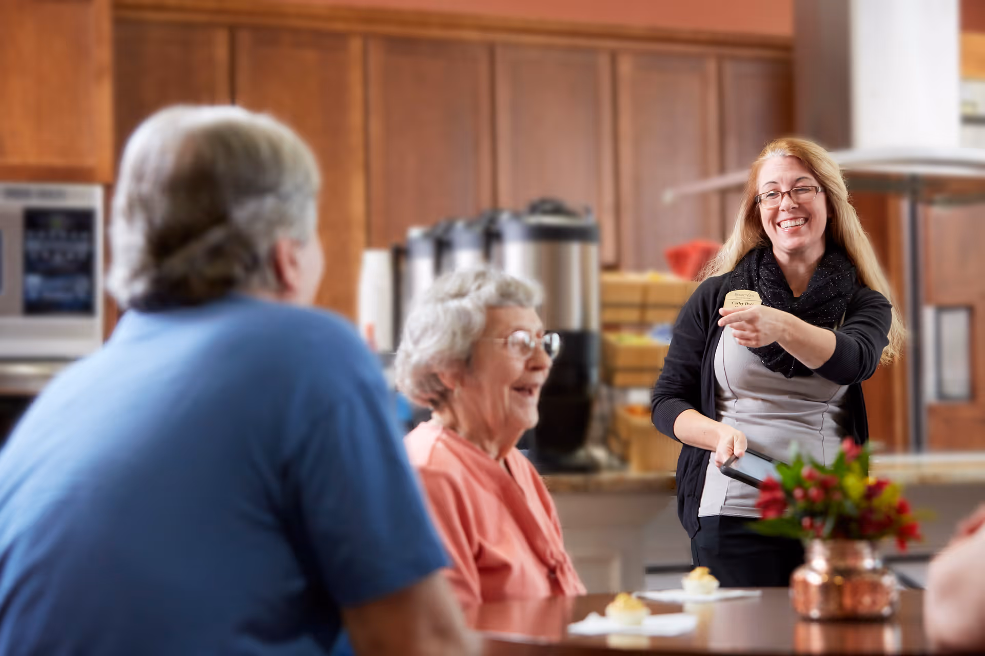 A smiling staff member with a name tag is serving food to elderly residents seated at a table in a warm, wood-paneled dining area with coffee dispensers in the background.