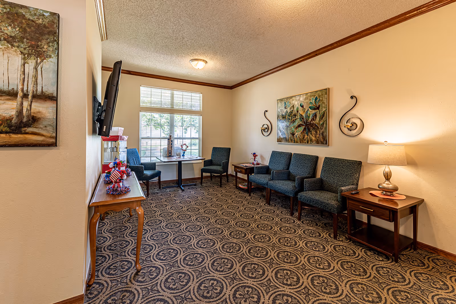 A cozy sitting area in Ashbrook Village with patterned carpet, several blue upholstered chairs, a small table with a lamp, wall-mounted TV, and decorative paintings on the walls. A window lets in natural light, and there are small patriotic decorations on a wooden console table.