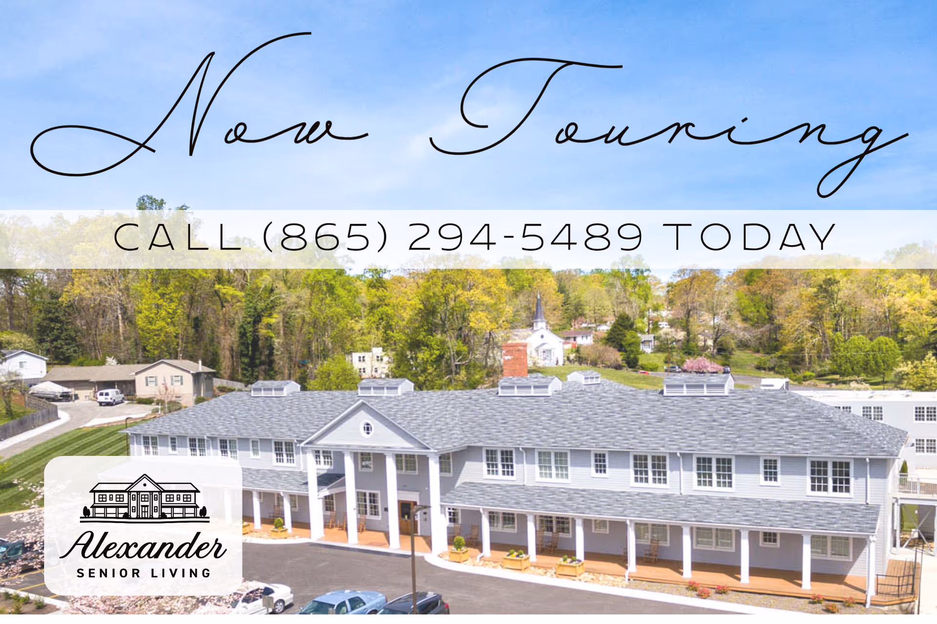 A large two-story senior living facility building with a gray roof and white exterior, surrounded by greenery and trees under a clear blue sky. There are cars parked in front of the building and a small church visible in the background.