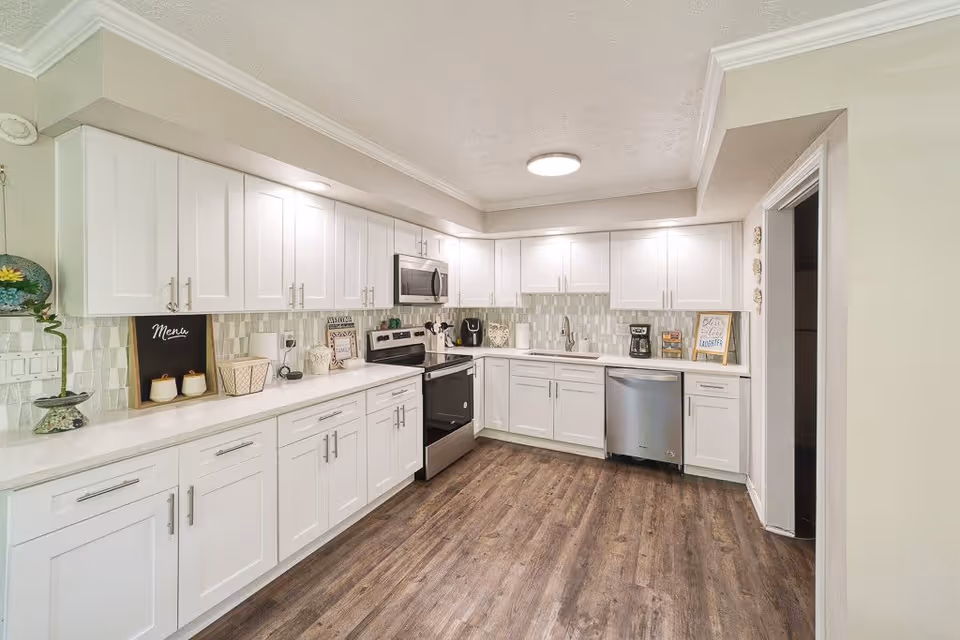 A modern kitchen with white cabinets, stainless steel appliances including a stove, microwave, and dishwasher. The kitchen has a light-colored backsplash, wooden flooring, and various decorative items on the countertops such as a small chalkboard menu, a coffee maker, and framed signs.