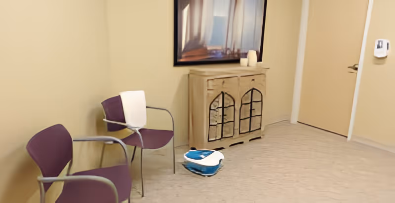 Small seating area with two purple chairs, a distressed wooden cabinet under a framed picture, and a blue-and-white device on the floor by a closed door.