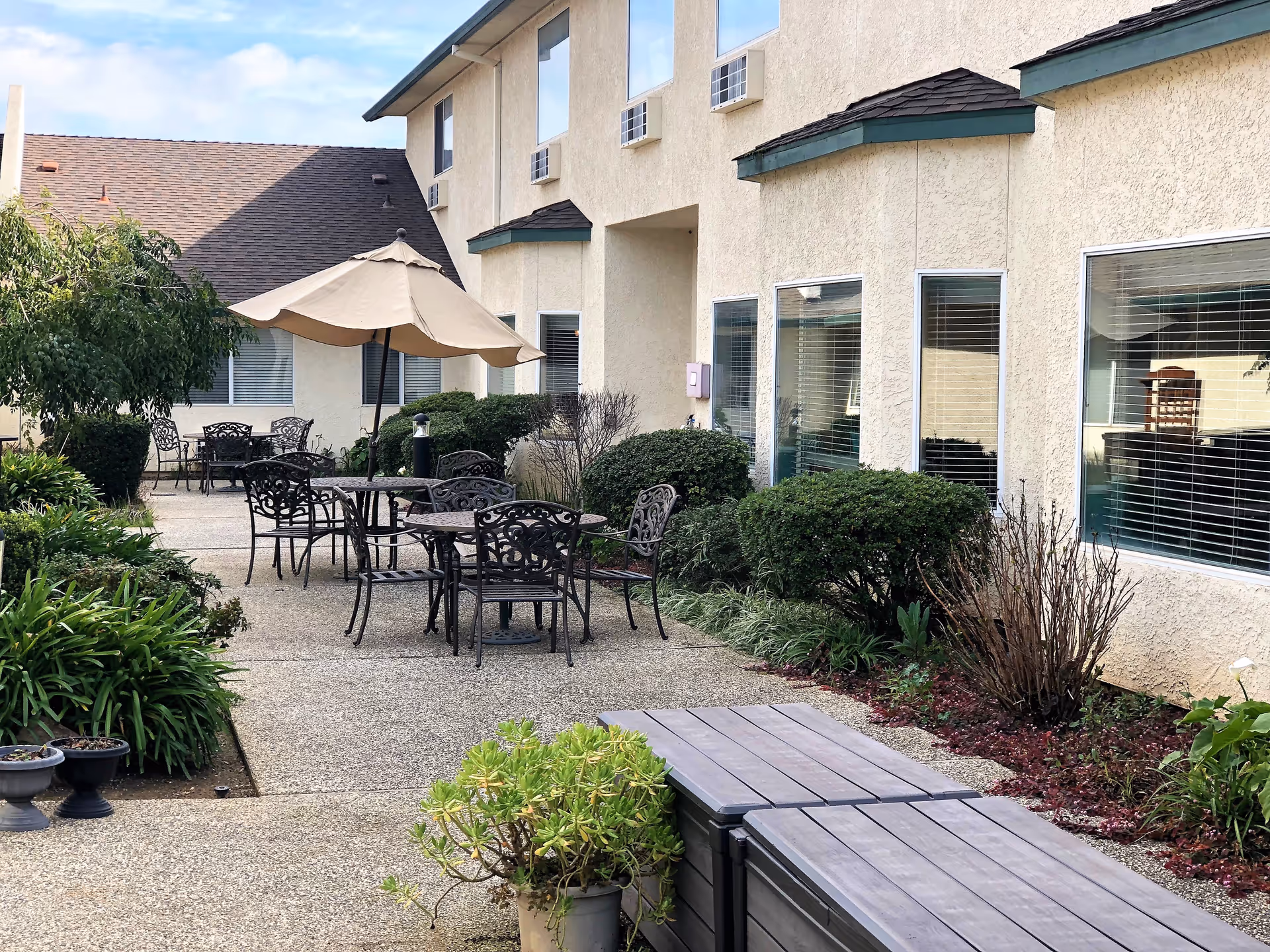 Sunny outdoor courtyard with metal tables and chairs, a large umbrella, potted plants, and a beige stucco building with windows.