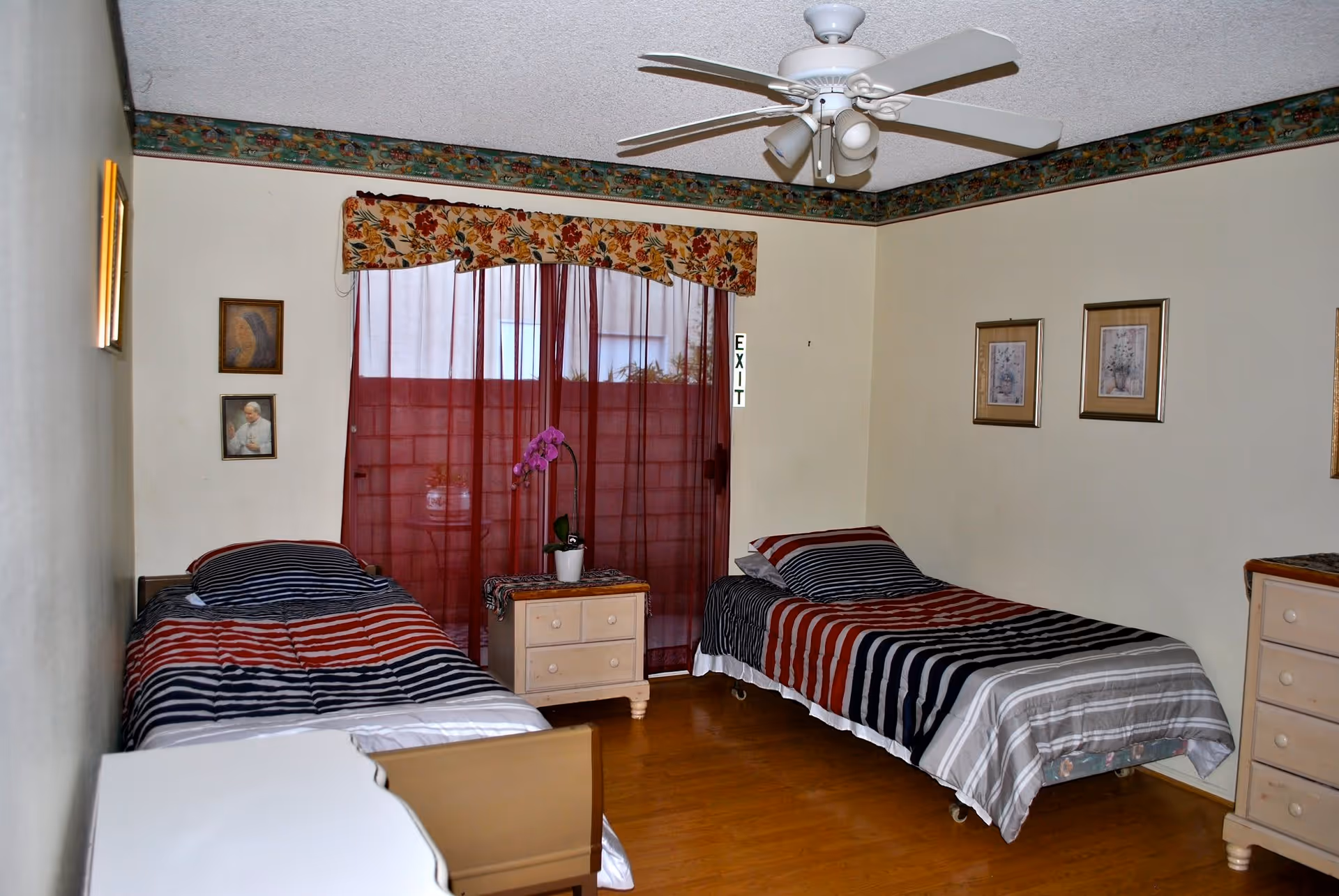 A bedroom with two single beds, each with striped bedding in shades of red, black, and gray. Between the beds is a small nightstand with a potted orchid on top. The room has light-colored walls with floral border wallpaper near the ceiling, a ceiling fan with lights, and framed pictures on the walls. A window with sheer red curtains and a floral valance is visible behind the nightstand.