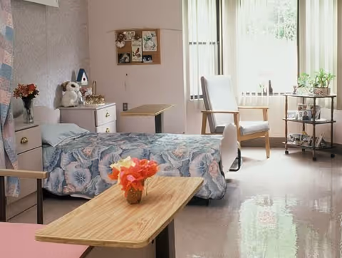 Sunlit resident bedroom with a single bed in a floral bedspread, an armchair by the window, bedside furniture, and a small vase of flowers on an overbed table.