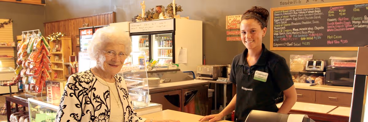 An elderly woman with white hair and glasses stands at a counter in a small café or deli area, smiling. Behind the counter is a young female employee wearing a black uniform and name tag, also smiling. The background shows a menu board with various food and drink options, a refrigerator with beverages, and shelves with snacks.