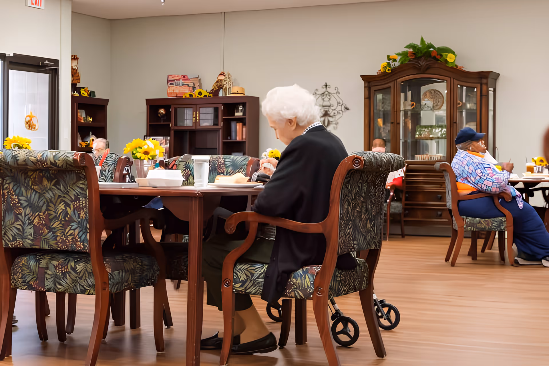 Elderly residents sitting at tables in a well-lit common dining area with wooden furniture, floral decorations, and a glass cabinet in the background.
