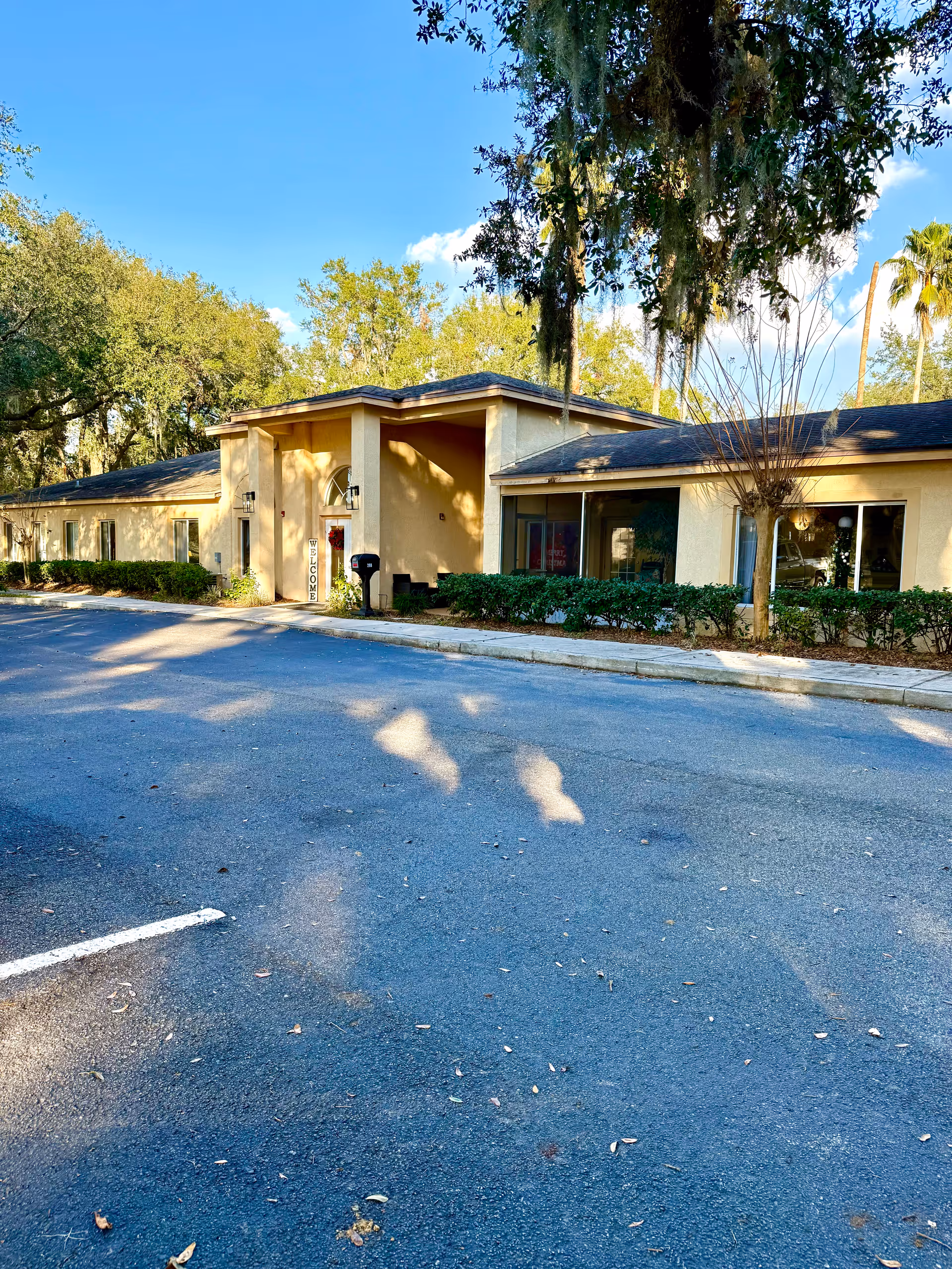 Exterior view of a single-story senior living facility building with beige walls and a dark roof, surrounded by trees and bushes under a clear blue sky. There is a parking lot in front of the building and a vertical 'WELCOME' sign near the entrance.