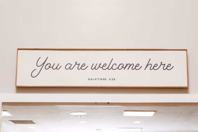 A rectangular wooden-framed sign mounted on a wall with the text 'You are welcome here' in cursive and 'GALATIANS 3:28' in smaller print below it.