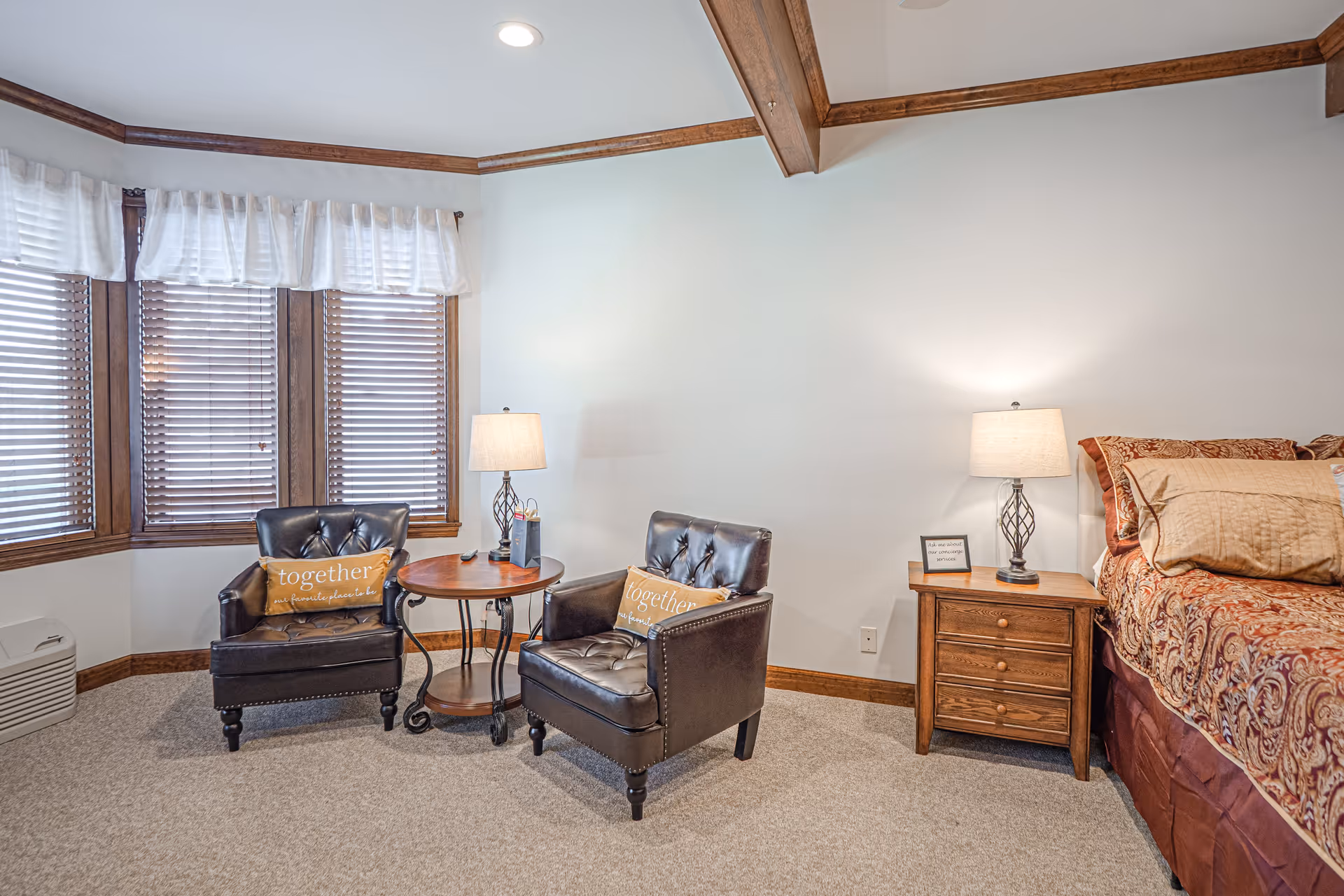 A cozy bedroom corner featuring two dark leather armchairs with decorative pillows that say 'together is our favorite place to be,' a round wooden side table between them, a wooden nightstand with a lamp and a small framed quote, and a bed with patterned bedding. The room has beige carpet, white walls, wooden trim, and a window with wooden blinds and white valances.
