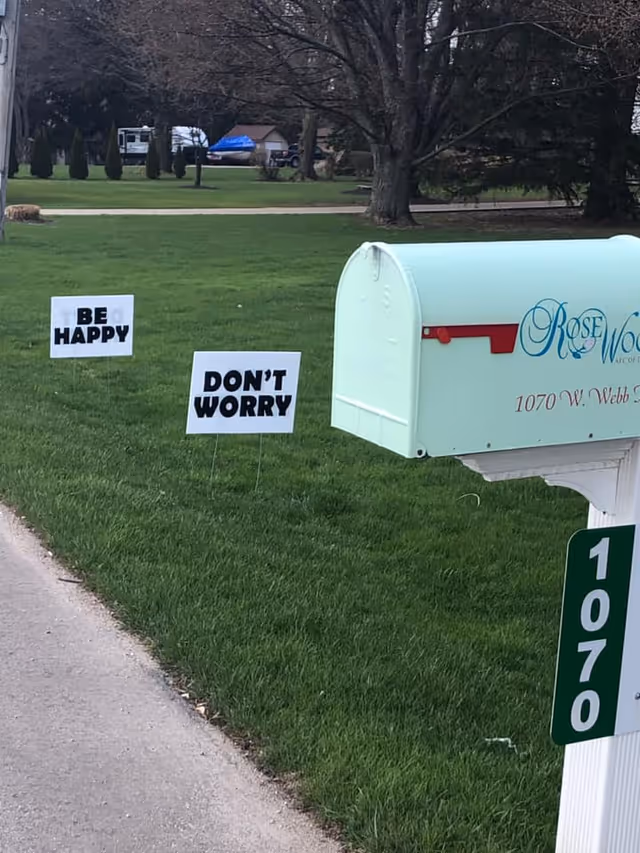 A light-green mailbox marked Rosewood and address 1070 sits on a grassy lawn with two yard signs reading "BE HAPPY" and "DON'T WORRY."