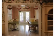 Interior view of a dining area with two tables and chairs on a tiled floor. The room has large glass doors with curtains and decorative valances above them. There is a wooden cabinet on the right side and floral wallpaper on the walls.