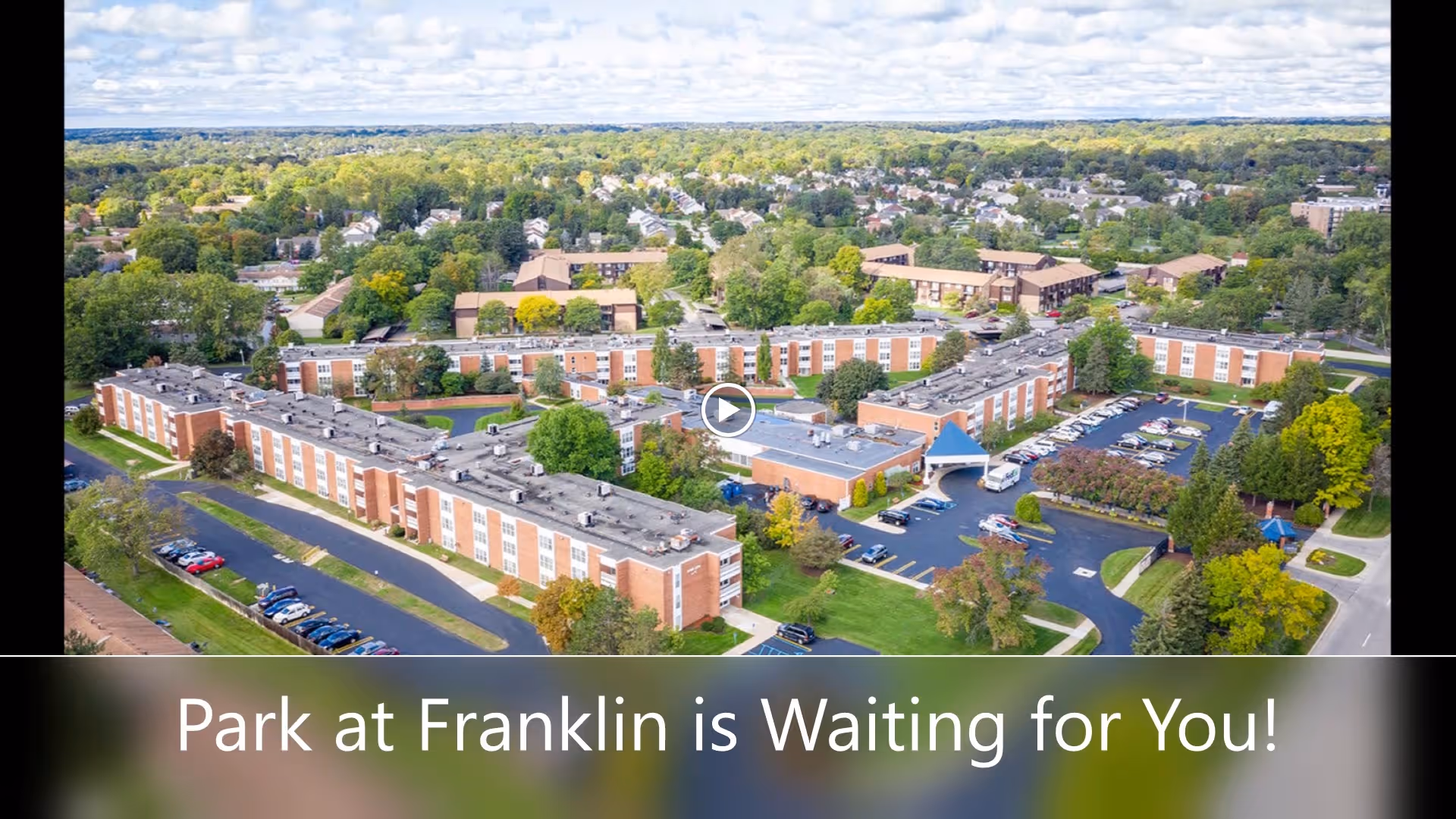 Aerial view of the Park At Franklin senior living facility showing multiple connected brick buildings surrounded by trees, parking lots, and roads under a partly cloudy sky.