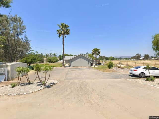View of a single-story building with a gray roof and walls, surrounded by a dirt and paved driveway. There are palm trees and other greenery around the building, a white car parked on the right side, and a clear blue sky overhead.