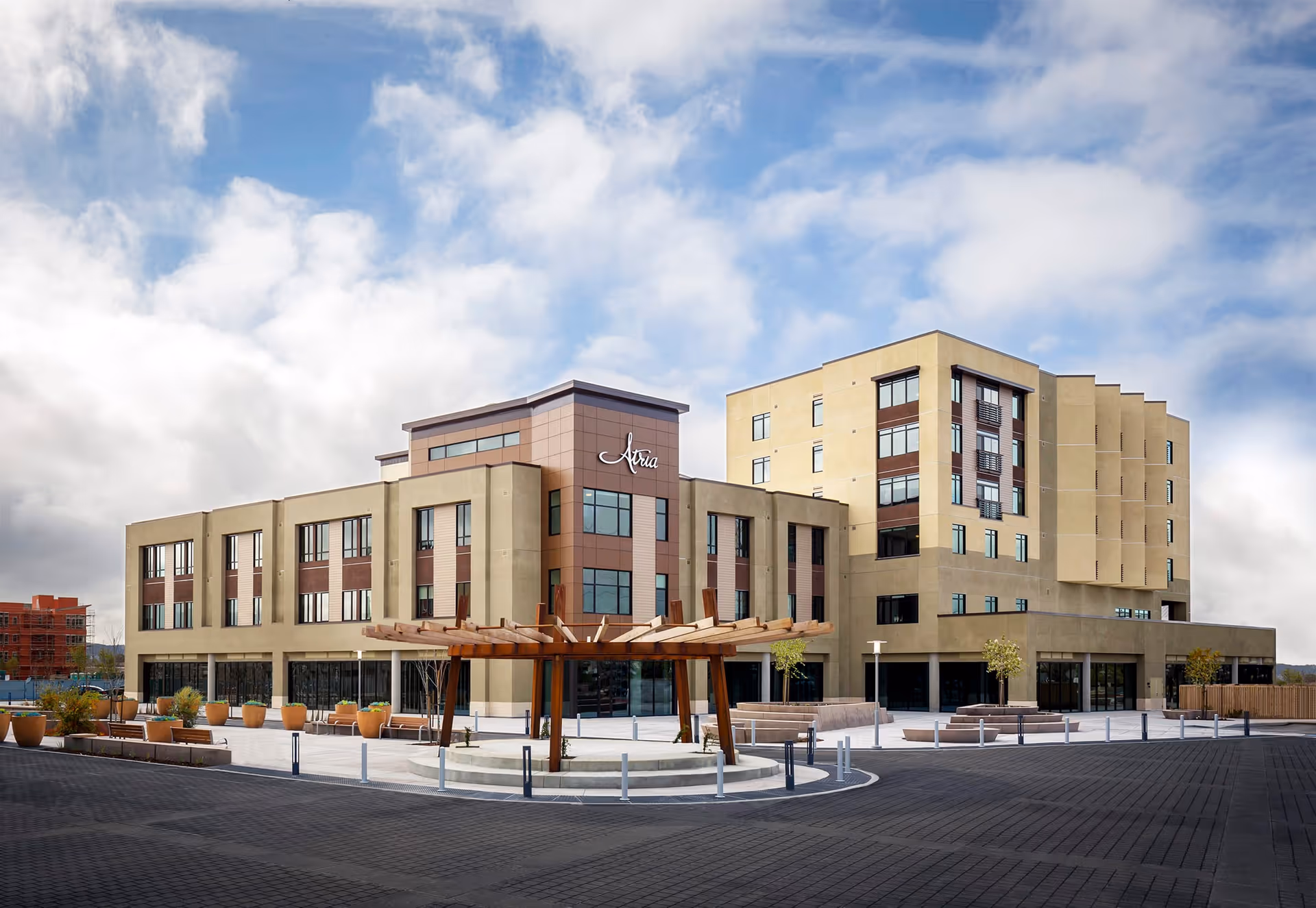 Exterior view of a modern multi-story senior living facility named Atria At Foster Square under a partly cloudy sky, featuring large windows, a wooden pergola structure, and landscaped planters in front.