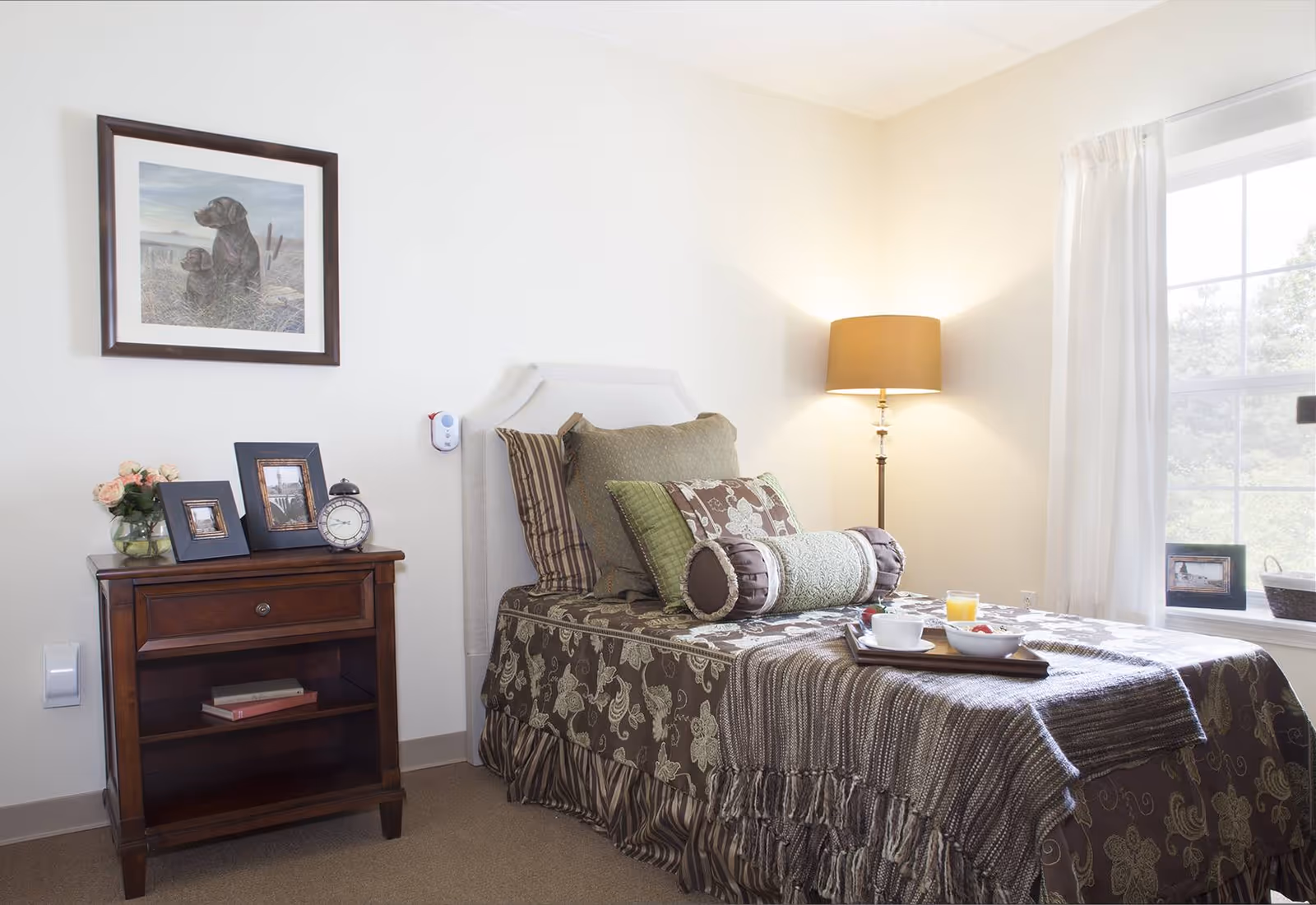 A neatly decorated single bedroom featuring a bed with patterned bedding, a wooden nightstand with framed photos and a clock, a floor lamp, and a window letting in natural light.