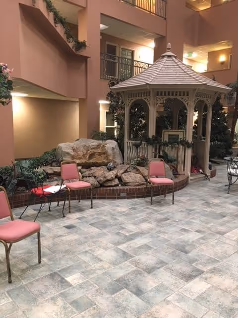 Indoor common area with a tiled floor, several pink cushioned chairs arranged around a small black metal table, a decorative rock water feature, and a white wooden gazebo. The area is surrounded by multiple floors with balconies and warm lighting.