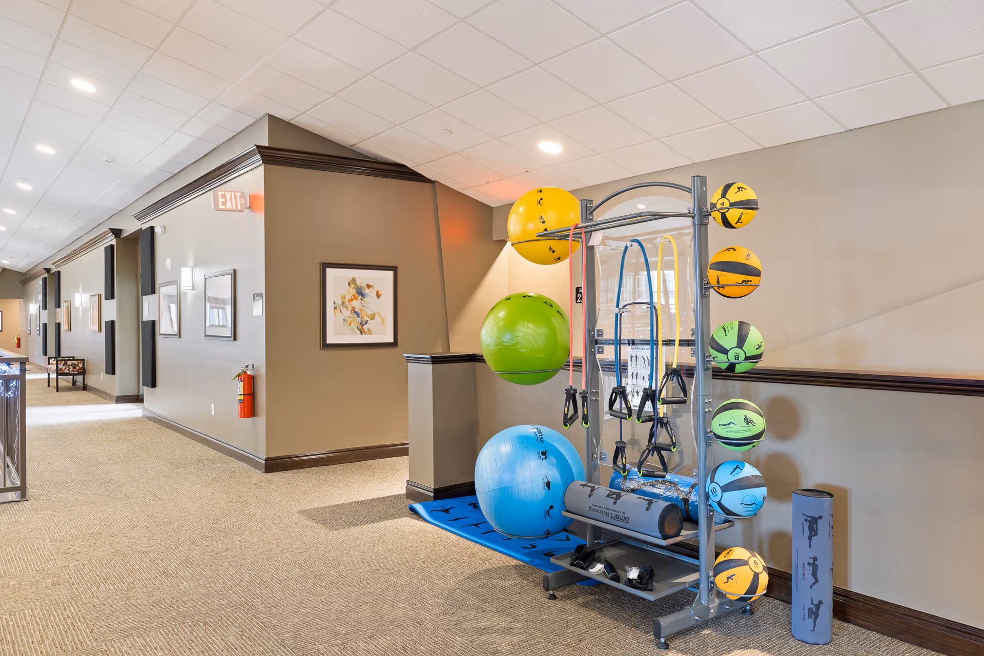 A hallway in a senior living facility with beige walls and carpeted floor. On the right side, there is a rack holding various exercise equipment including colorful stability balls, resistance bands, and foam rollers. The hallway is well-lit with ceiling lights and has framed artwork on the walls. An exit sign and a fire extinguisher are visible on the left wall.