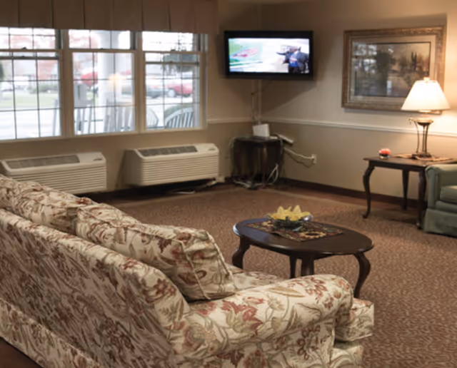 A cozy living room with a floral patterned sofa facing a small wooden coffee table. A flat-screen TV is mounted on the wall above a small table with electronic devices. Two large windows with blinds let in natural light. A side table with a lamp and a framed picture on the wall add to the warm ambiance.