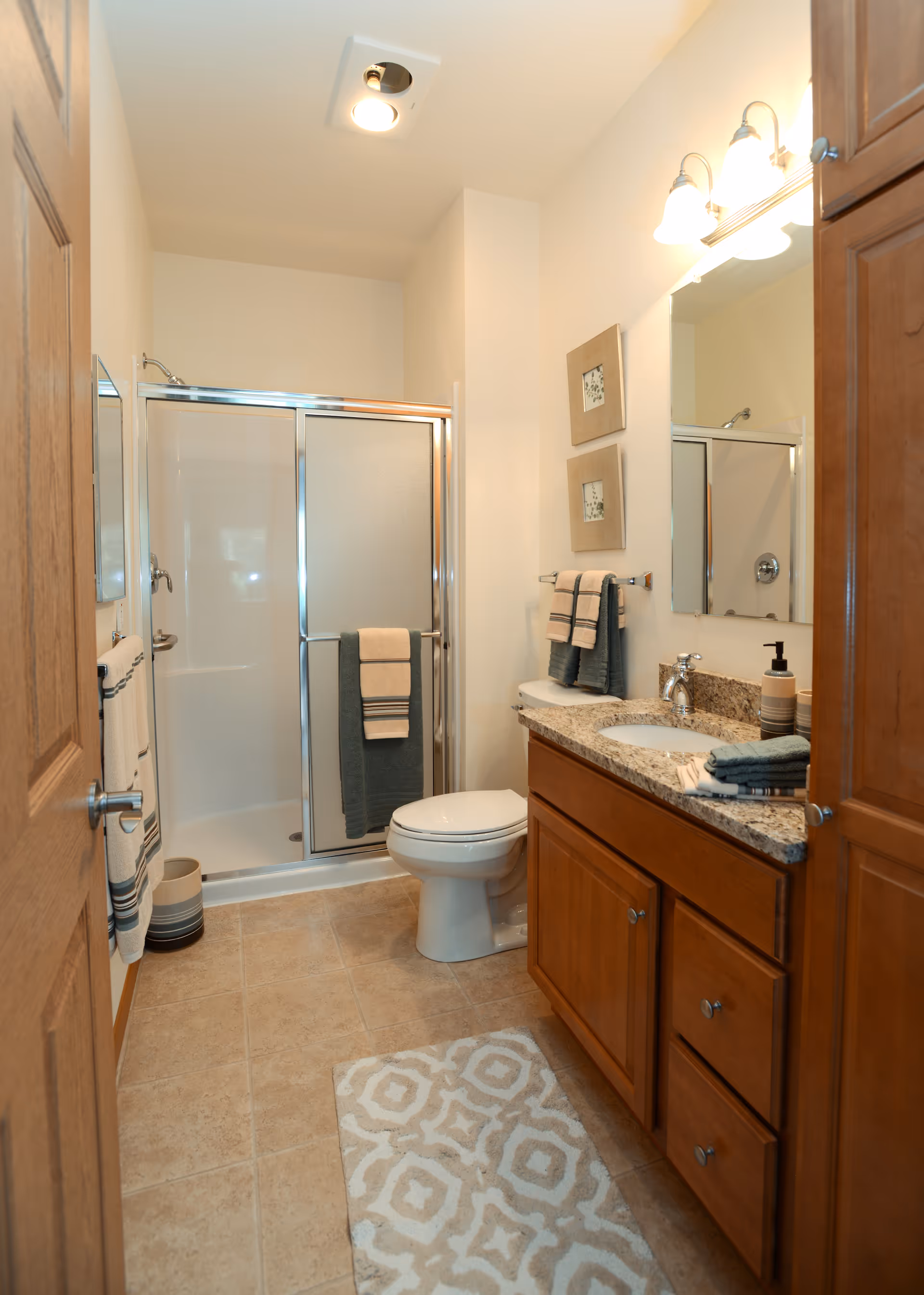 A clean and well-lit bathroom featuring a shower with sliding glass doors, a toilet, and a wooden vanity with a granite countertop. The vanity has a sink, soap dispenser, and neatly folded towels. The walls are decorated with two framed pictures, and there are multiple towels hanging on racks. The floor is tiled and has a patterned bath mat.