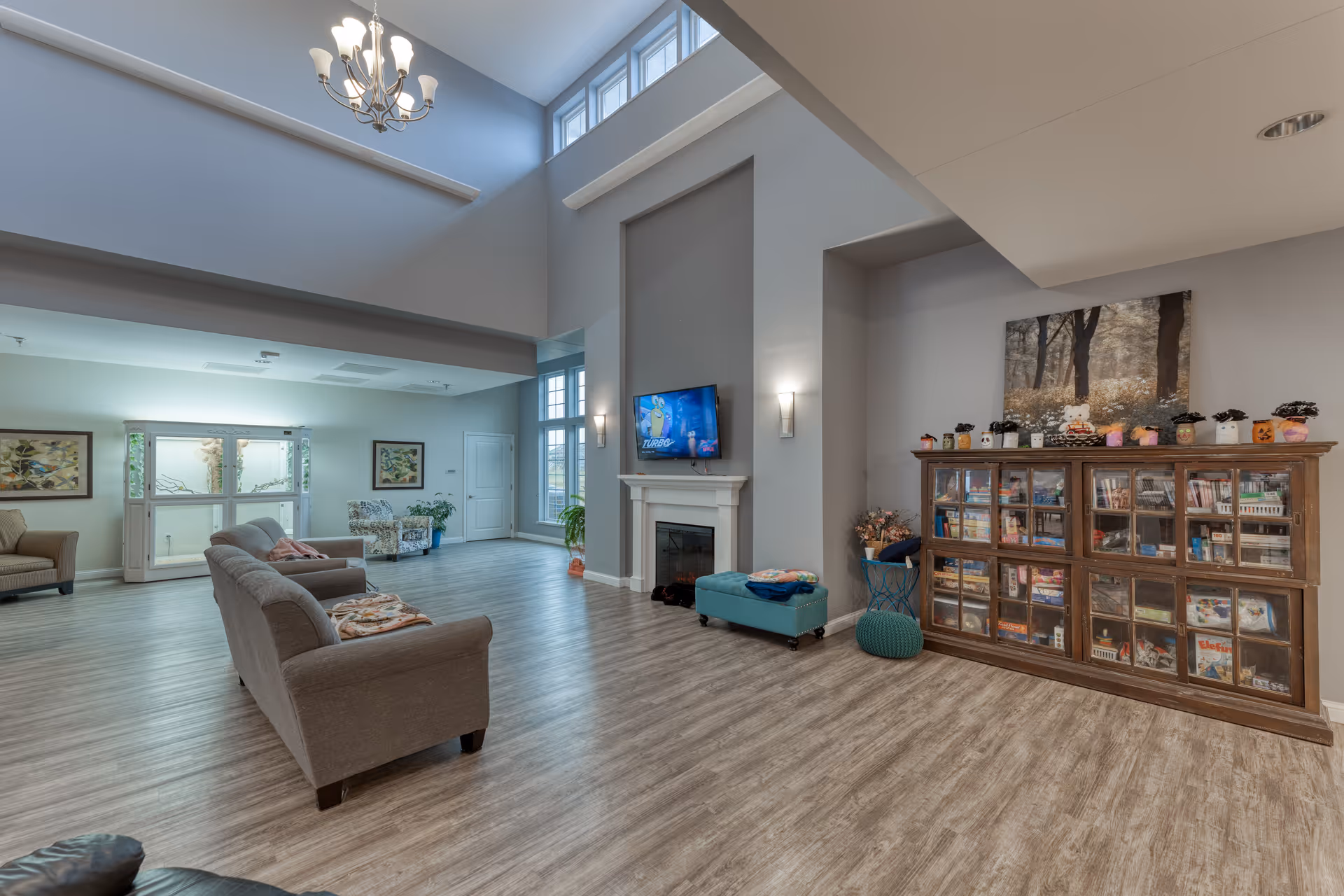 Spacious senior living common room with sofas, a fireplace and TV, a large bookcase, and high ceilings with chandelier lighting.
