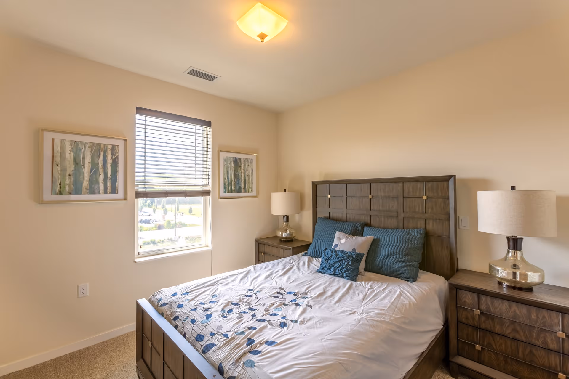A bedroom with a large wooden bed featuring a headboard and white bedding with blue floral patterns. There are three pillows on the bed in shades of blue and gray. On each side of the bed are matching wooden nightstands with modern table lamps. The room has beige walls, a window with blinds, and two framed pictures of trees hanging on the wall beside the window.