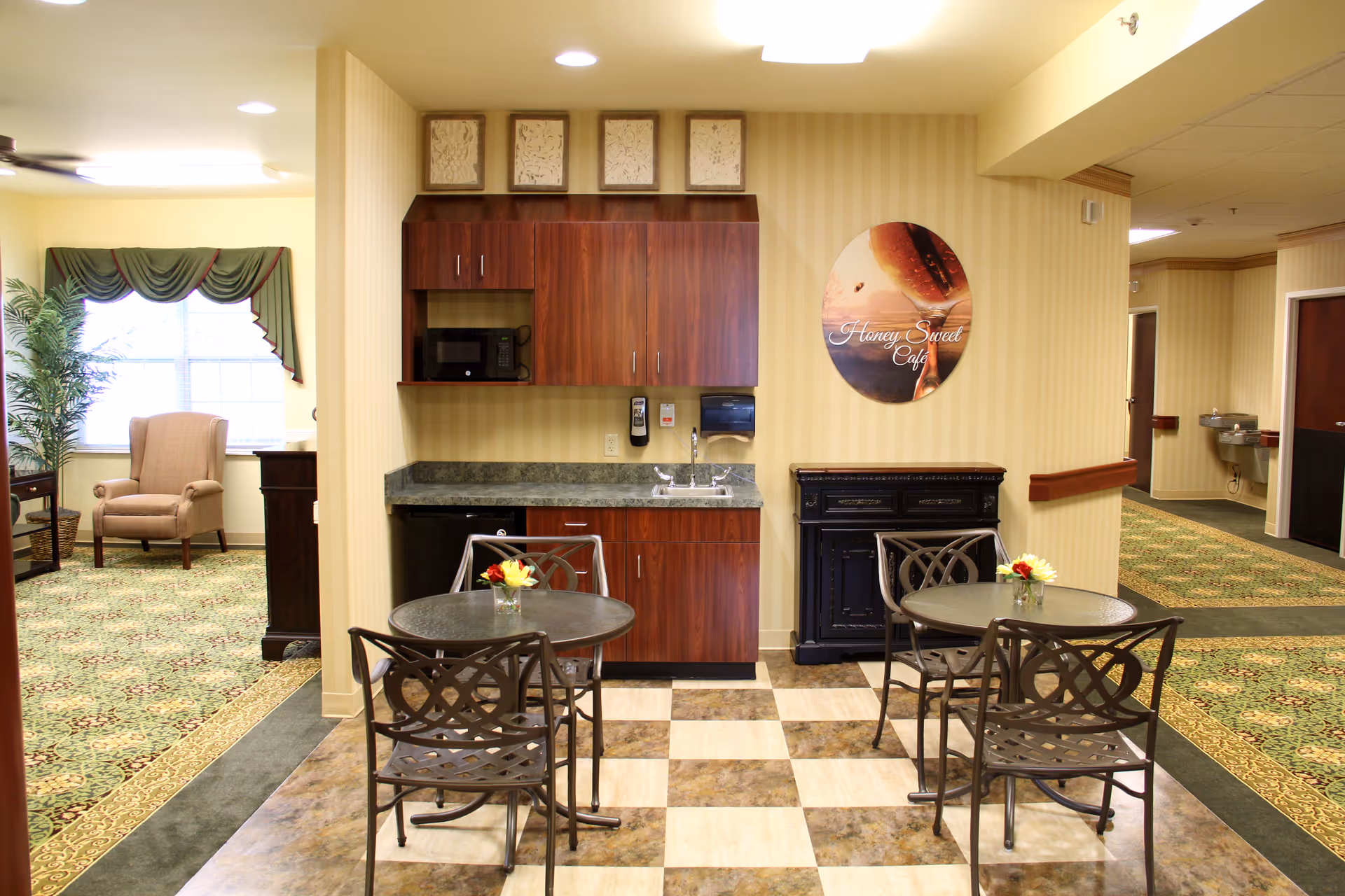 A small cafe area inside a senior living facility featuring two round tables with metal chairs, each table decorated with a small vase of flowers. The back wall has wooden cabinets, a microwave, a sink, and a paper towel dispenser. A circular sign on the wall reads 'Honey Sweet Cafe'. To the left, there is a sitting area with an armchair near a window with green curtains and a tall potted plant. The floor has a checkered tile pattern in the cafe area and patterned carpet in the sitting area and hallway.
