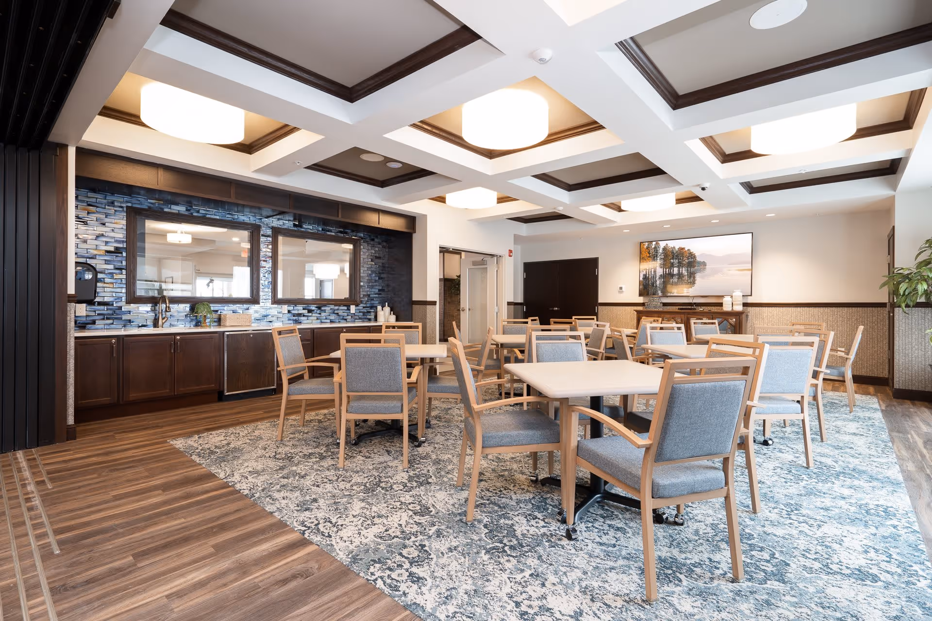 A bright and spacious dining room with multiple square tables and wooden chairs with gray cushions arranged on a patterned blue and white carpet. The room features a coffered ceiling with modern light fixtures, a blue tiled backsplash with two large mirrors above a countertop and cabinets, and a large landscape painting on the far wall. The floor is a combination of wood and carpet, and there is a plant in the corner.