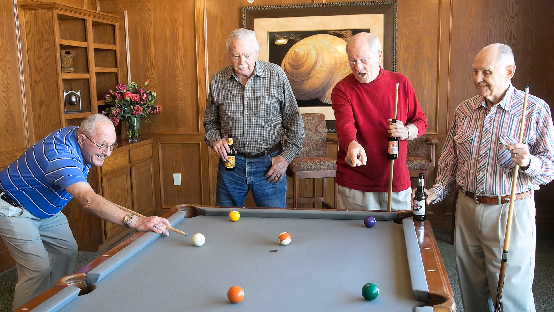 Four elderly men playing pool in a wood-paneled room. One man is taking a shot while the others watch, holding pool cues and bottles of beer. There is a framed picture on the wall and a vase with flowers on a shelf in the background.