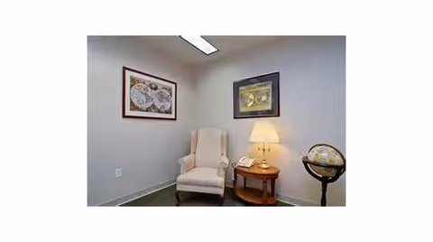 A small corner of a room with a beige upholstered armchair next to a wooden side table holding a lamp and a telephone. Two framed pictures hang on the light-colored walls, and a globe stands on a black stand to the right of the table.