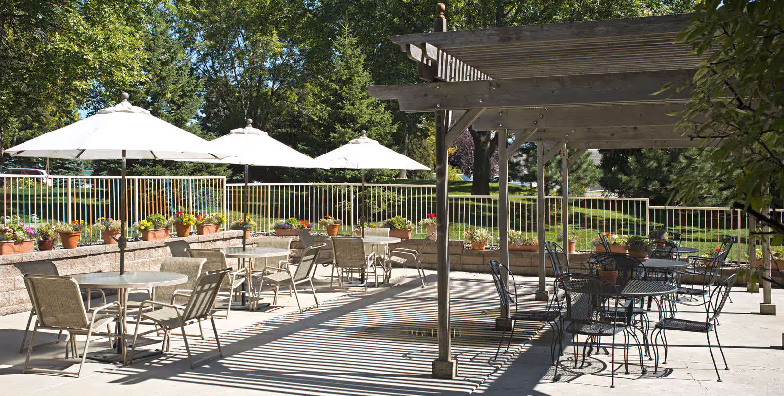 Outdoor patio area with multiple tables and chairs, some shaded by large white umbrellas and others under a wooden pergola. The patio is surrounded by a low brick wall with potted flowers and greenery, with trees and grass visible in the background.